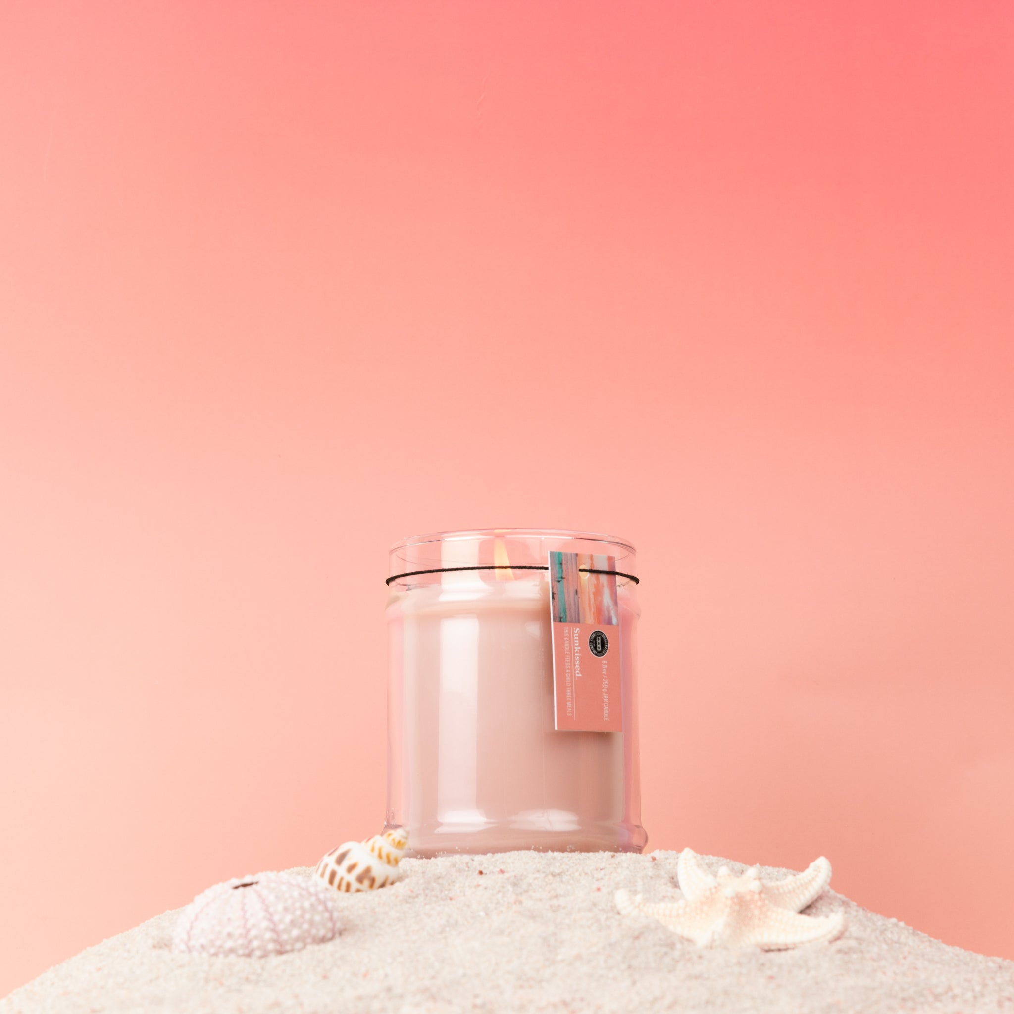 The Sunkissed 8oz Jar Candle by Bridgewater Candle, made with soy-blend wax, sits on sand amid seashells and a starfish against a soft pink gradient. A tag with blue and pink details hangs from the clear glass jar.