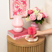 A round wooden side table holds a pink-striped lamp, pink book, and pink flowers. A hand sets a Bridgewater Candle Sweet Grace Honeycomb Diffuser on a pink-and-white coaster. A floral artwork is displayed on the wall.