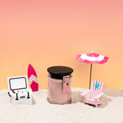 The Bridgewater Candle Sunkissed 8oz Jar Candle, made with soy-blend wax, sits on sand next to a pink surfboard, YETI cooler, striped beach umbrella, and beach chair amid a dreamy pink and orange gradient background.