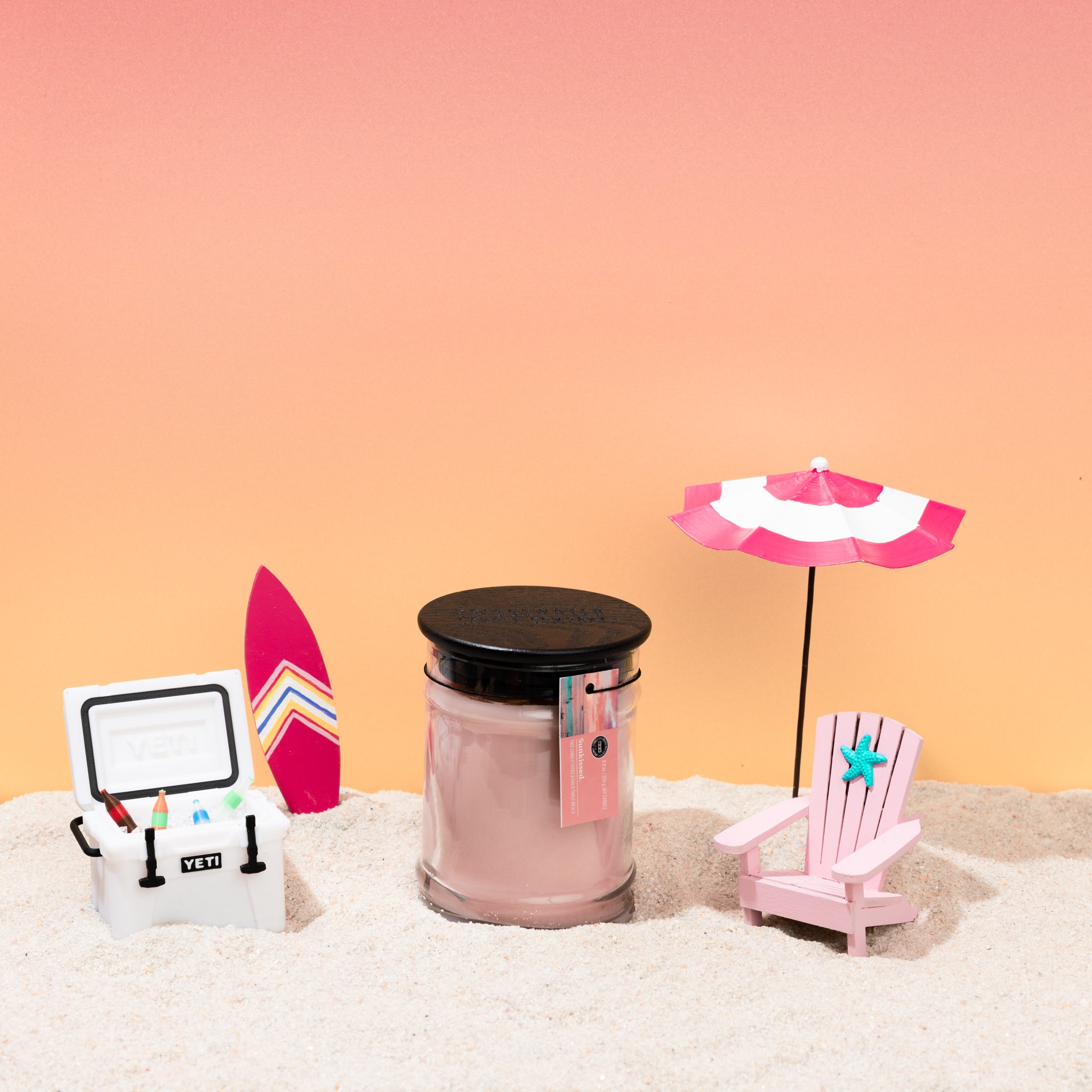 The Bridgewater Candle Sunkissed 8oz Jar Candle, made with soy-blend wax, sits on sand next to a pink surfboard, YETI cooler, striped beach umbrella, and beach chair amid a dreamy pink and orange gradient background.