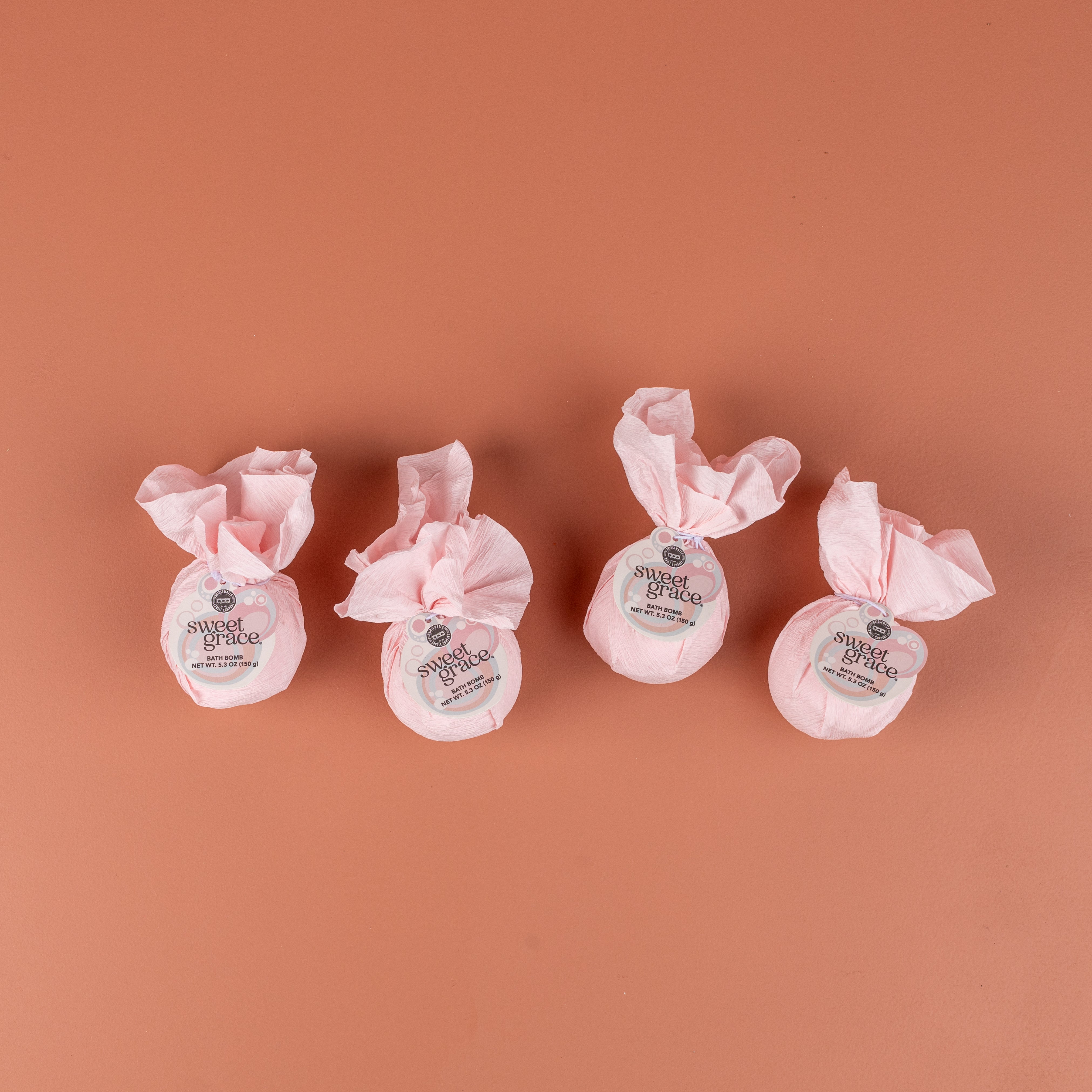 The Bath Bomb 4 Pack-Sweet Grace by Bridgewater Candle features four round bath bombs, wrapped in pink tissue with white labels, arranged side by side on a salmon-colored background.