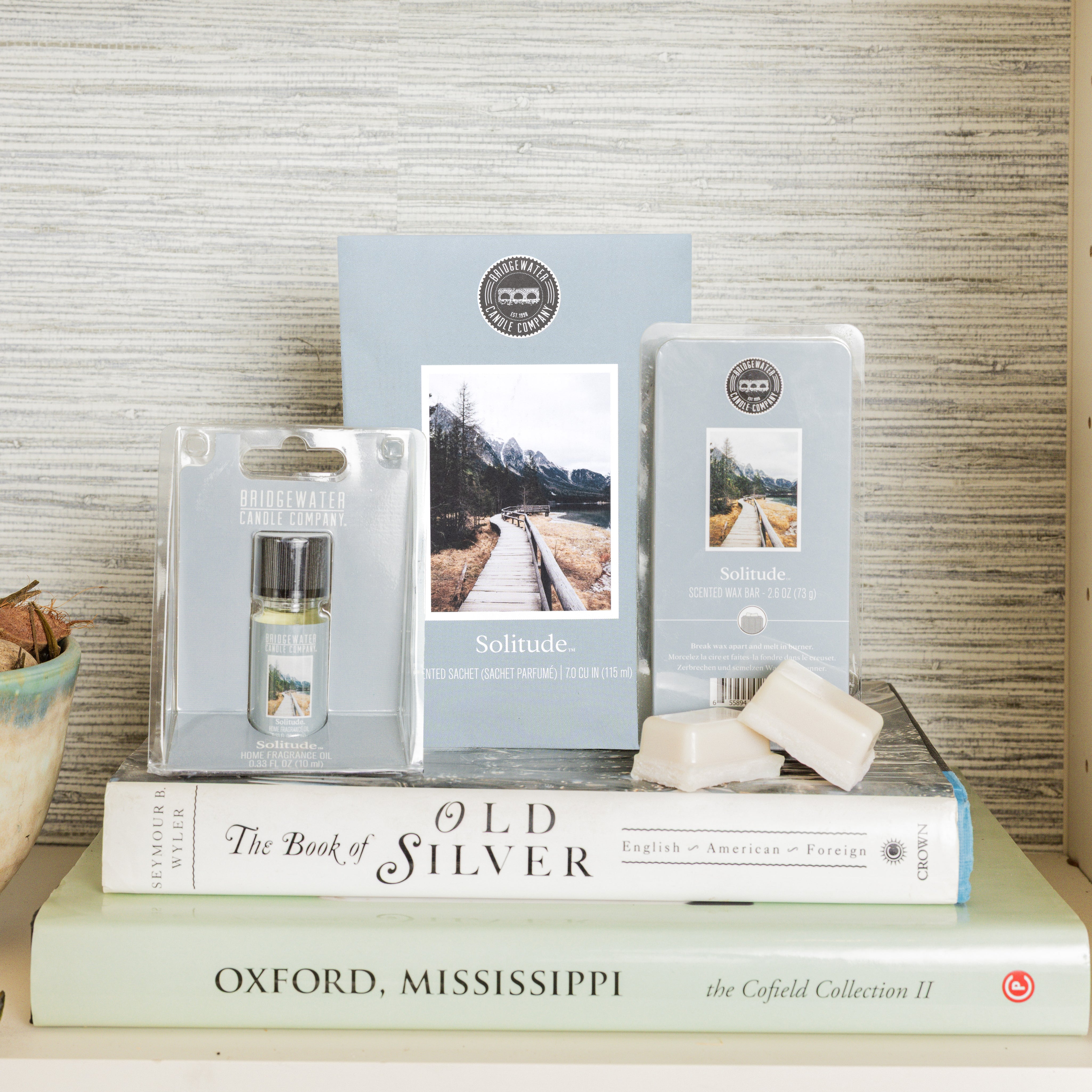 The Bridgewater Candle Solitude Flameless Bundle, featuring a small candle, wax melts, and home fragrance oil, is displayed on books including The Book of Old Silver and Oxford, Mississippi. A blue bowl rests beside the stack.