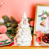 The Sweet Grace Collection Candle #70 by Bridgewater Candle, made from soy-blend wax, sits on a gold tray among festive décor, adding a cozy glow against a pink wall with holiday napkins, ornaments, greenery, and a framed bell illustration.