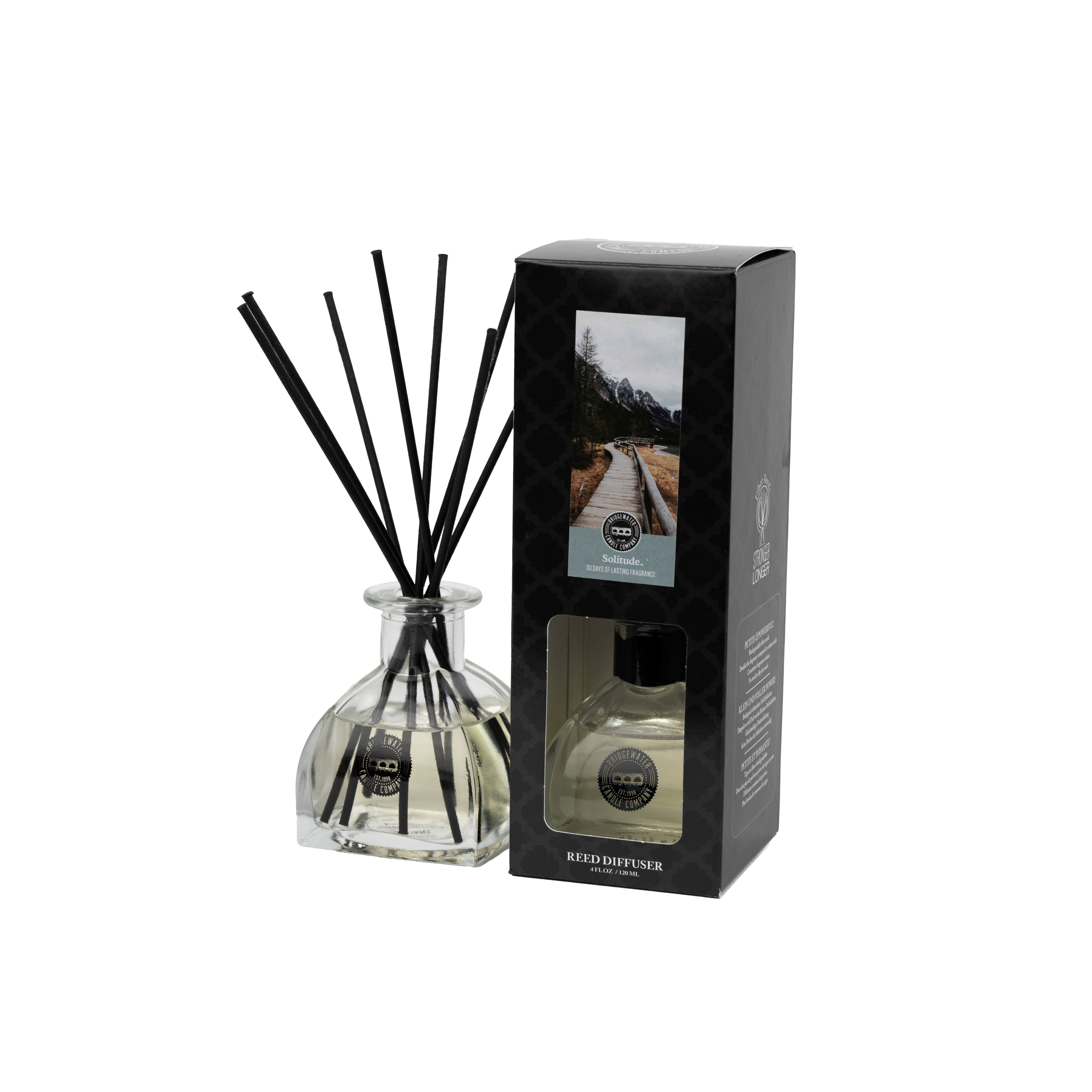 The Solitude Reed Diffuser by Bridgewater Candle features a clear glass bottle with black fiber reeds next to its black box packaging, which displays a mountain road image and product details. The diffuser holds a transparent premium home fragrance liquid.