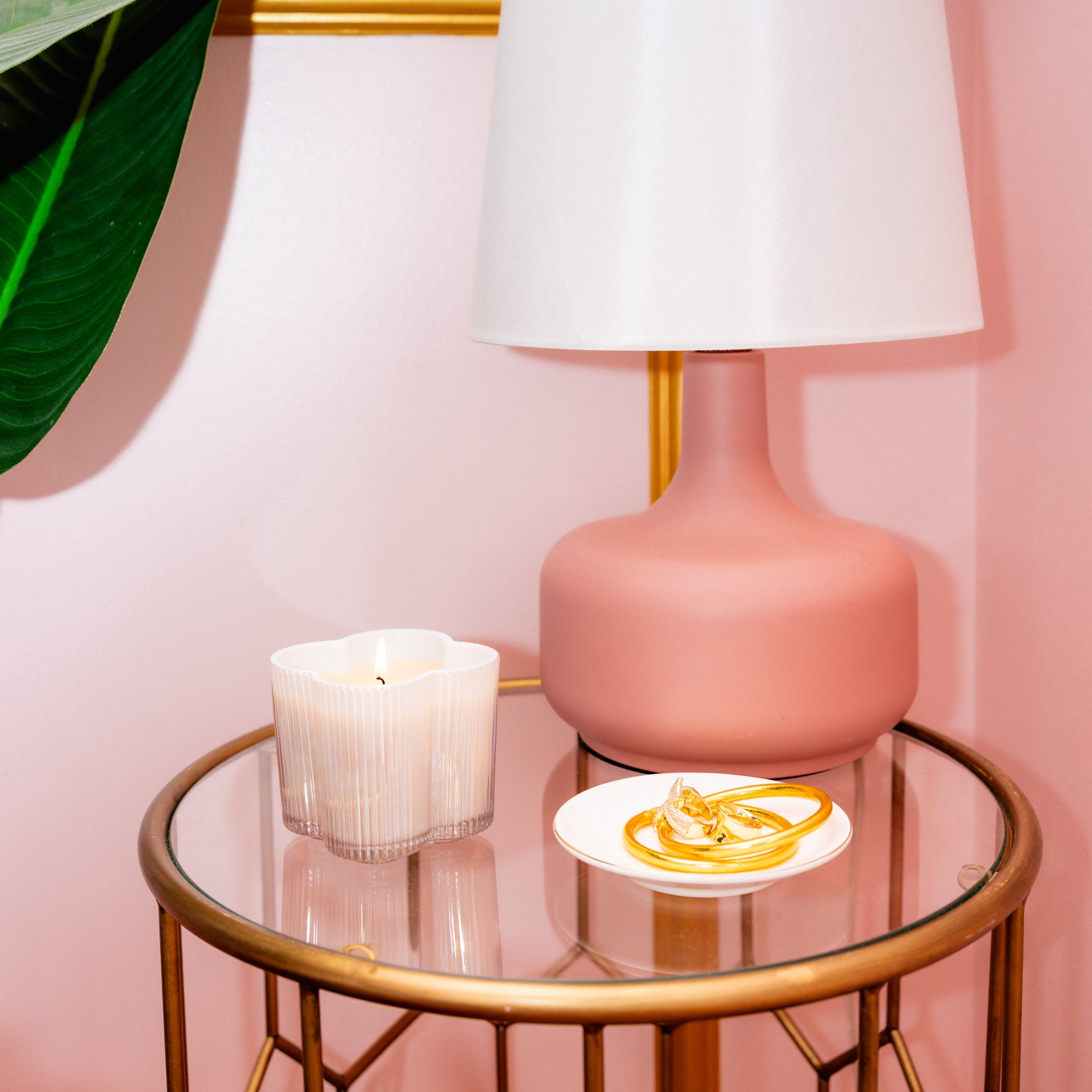 A round glass side table with a pink lamp, the lit Bridgewater Candle Sweet Grace Collection Candle #076 made from soy-blend wax, and a white dish with gold jewelry, sits against a pink wall with a large green leaf on the left.
