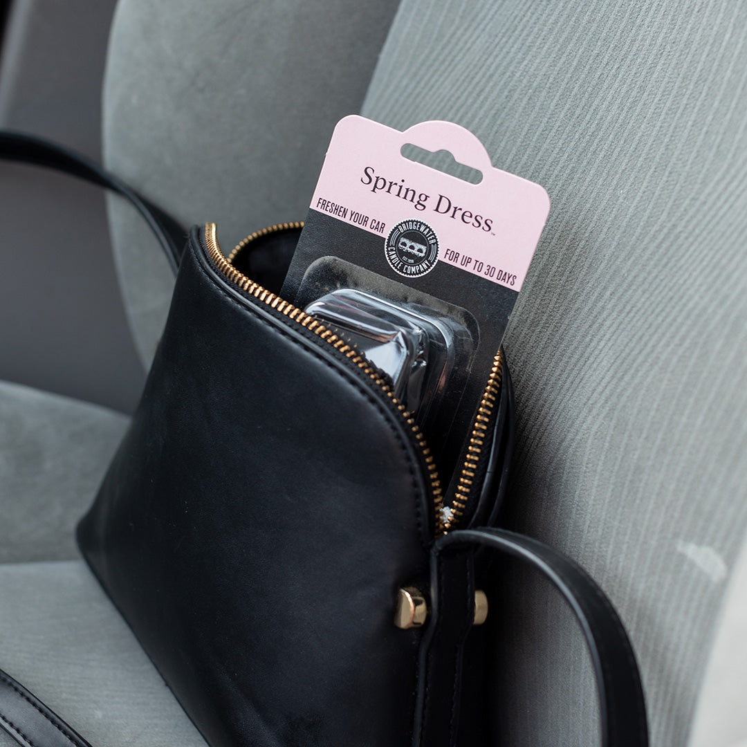 A Bridgewater Candle Auto Vent Clip-Spring Dress package tucked inside a small black purse resting on a car seat.
