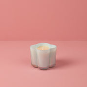 The Bridgewater Candle Sweet Grace Collection Candle #077, a white flower-shaped soy-blend wax candle in a ribbed glass container, is displayed on a pink surface against a solid pink background.