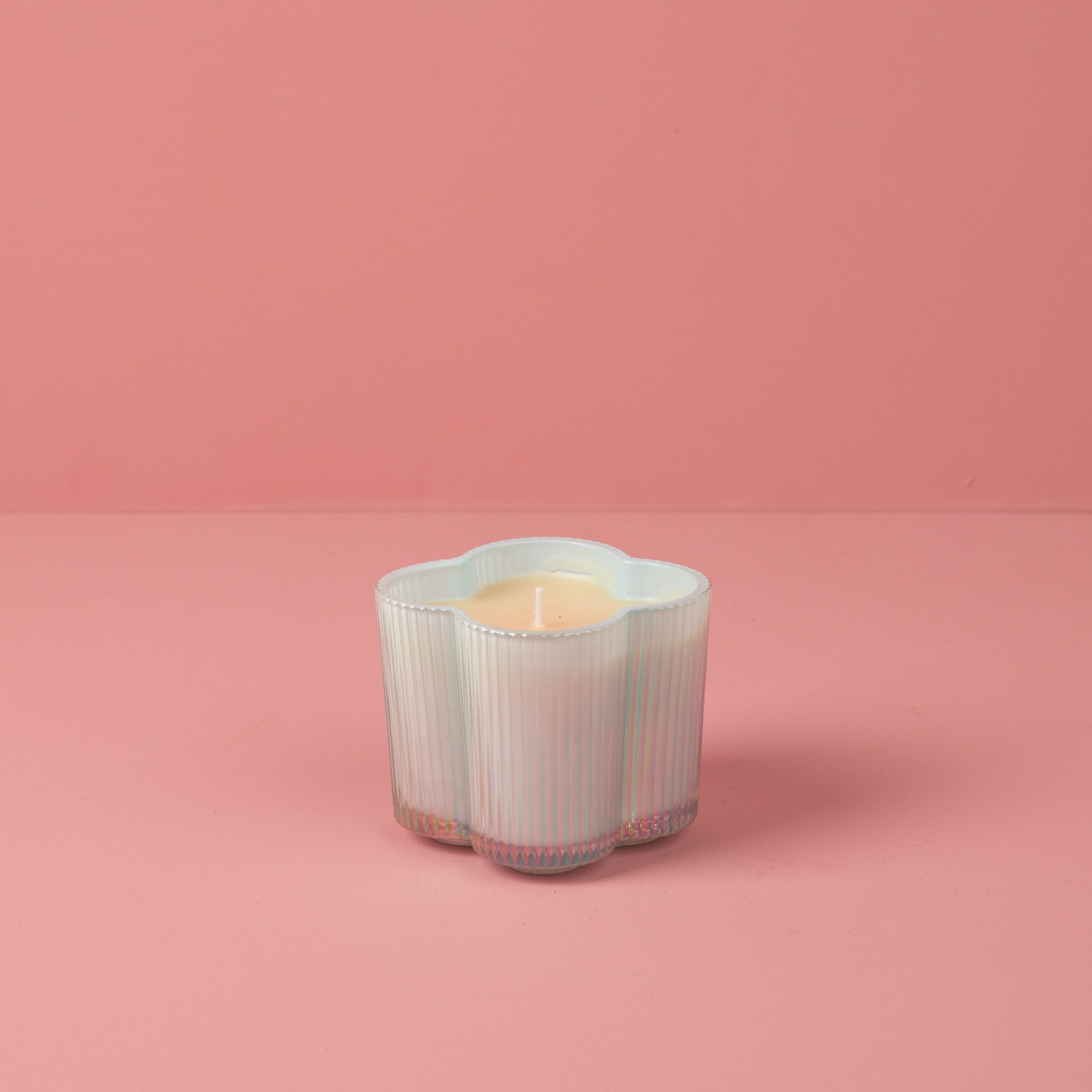 The Bridgewater Candle Sweet Grace Collection Candle #077, a white flower-shaped soy-blend wax candle in a ribbed glass container, is displayed on a pink surface against a solid pink background.