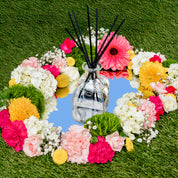 A clear High Bloom Reed Diffuser by Bridgewater Candle sits on a round mirror, surrounded by pink, yellow, and white flowers atop bright green grass—a beautiful touch of home décor.