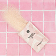 A pouch of Bridgewater Candle Sweet Grace Soaking Salts with Pink Himalayan sea salt rests on pink tiles, some salts spilled out to create textured contrast against the smooth surface.