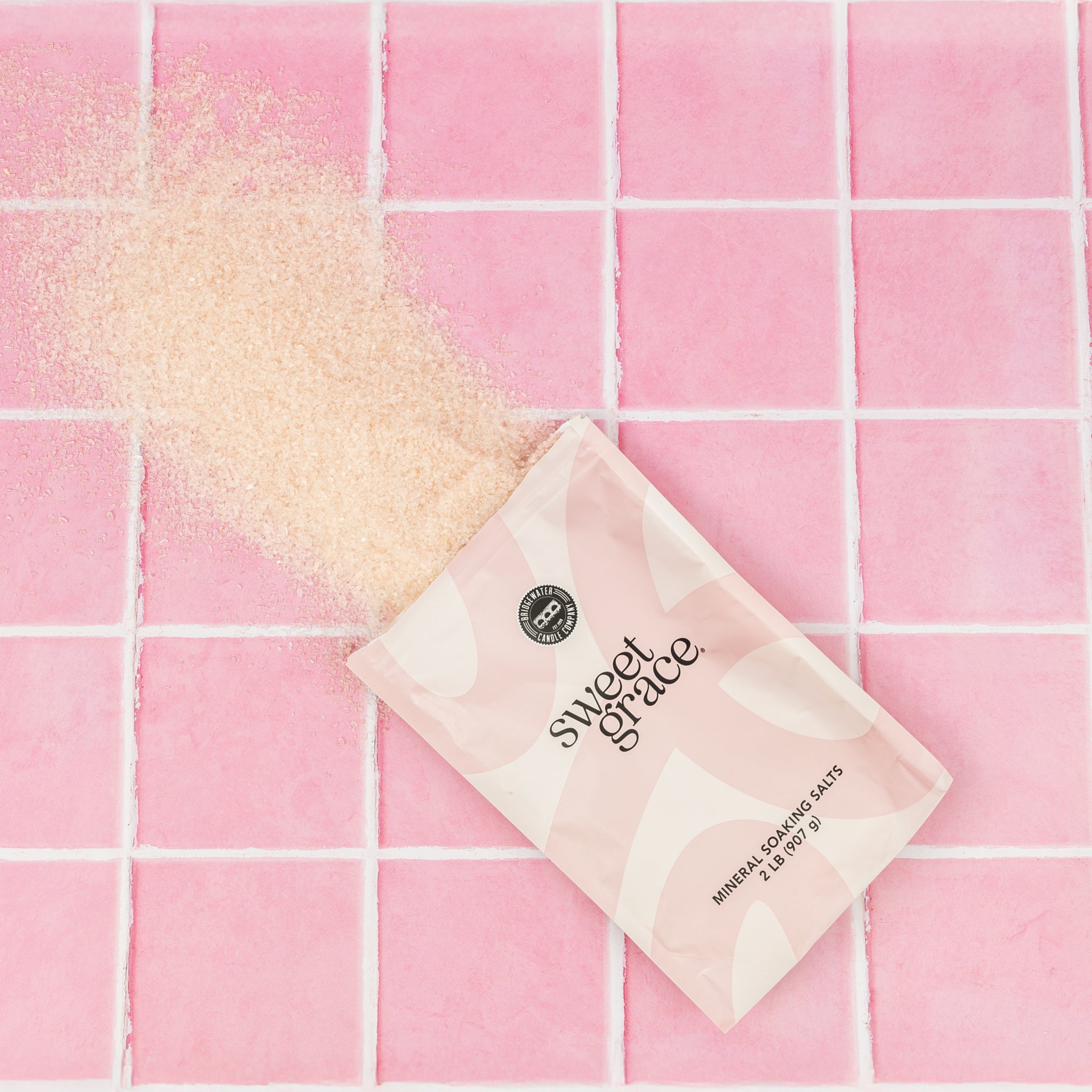 A pouch of Bridgewater Candle Sweet Grace Soaking Salts with Pink Himalayan sea salt rests on pink tiles, some salts spilled out to create textured contrast against the smooth surface.