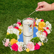 A hand lights the Bridgewater Candle High Bloom 18oz Large Jar Candle, made with soy-blend wax, set on a mirror and encircled by a colorful ring of flowers on green grass.