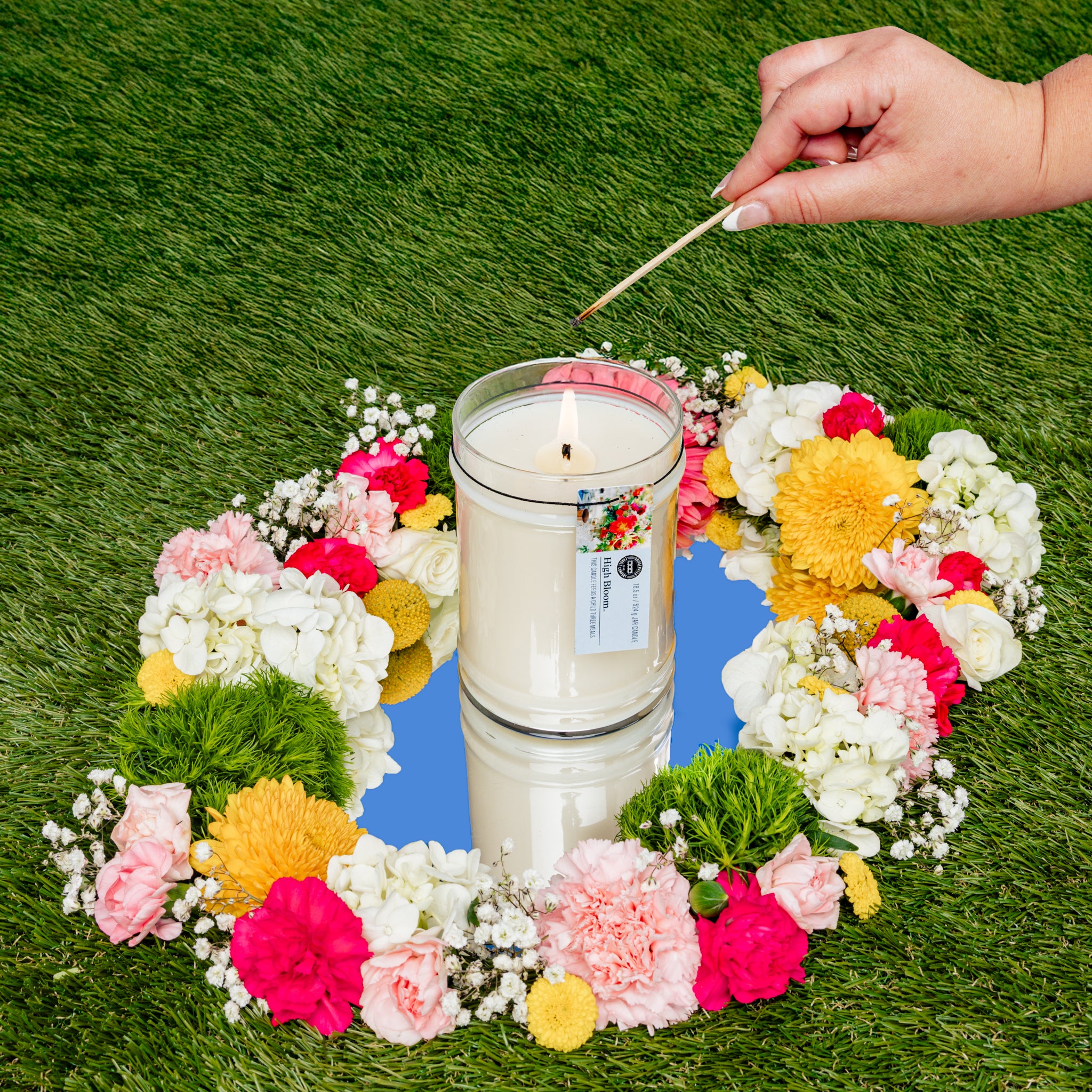 A hand lights the Bridgewater Candle High Bloom 18oz Large Jar Candle, made with soy-blend wax, set on a mirror and encircled by a colorful ring of flowers on green grass.
