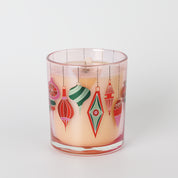 The Bridgewater Candle Sweet Grace Ornament Decal Candle features a glass holder filled with light peach, soy-blend wax and is adorned with colorful, retro-style holiday ornament illustrations in red, green, and pink on the outside.