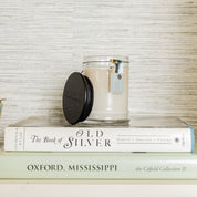 The Solitude Small Jar Candle by Bridgewater Candle, featuring soy-blend wax and a black lid, sits atop The Book of Old Silver and Oxford, Mississippi, against a textured background, filling your space with its signature scent.