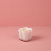 The Sweet Grace Collection Candle #076 by Bridgewater Candle is a small, white, flower-shaped soy-blend candle in a ribbed glass holder displayed on a pink surface against a pink background.