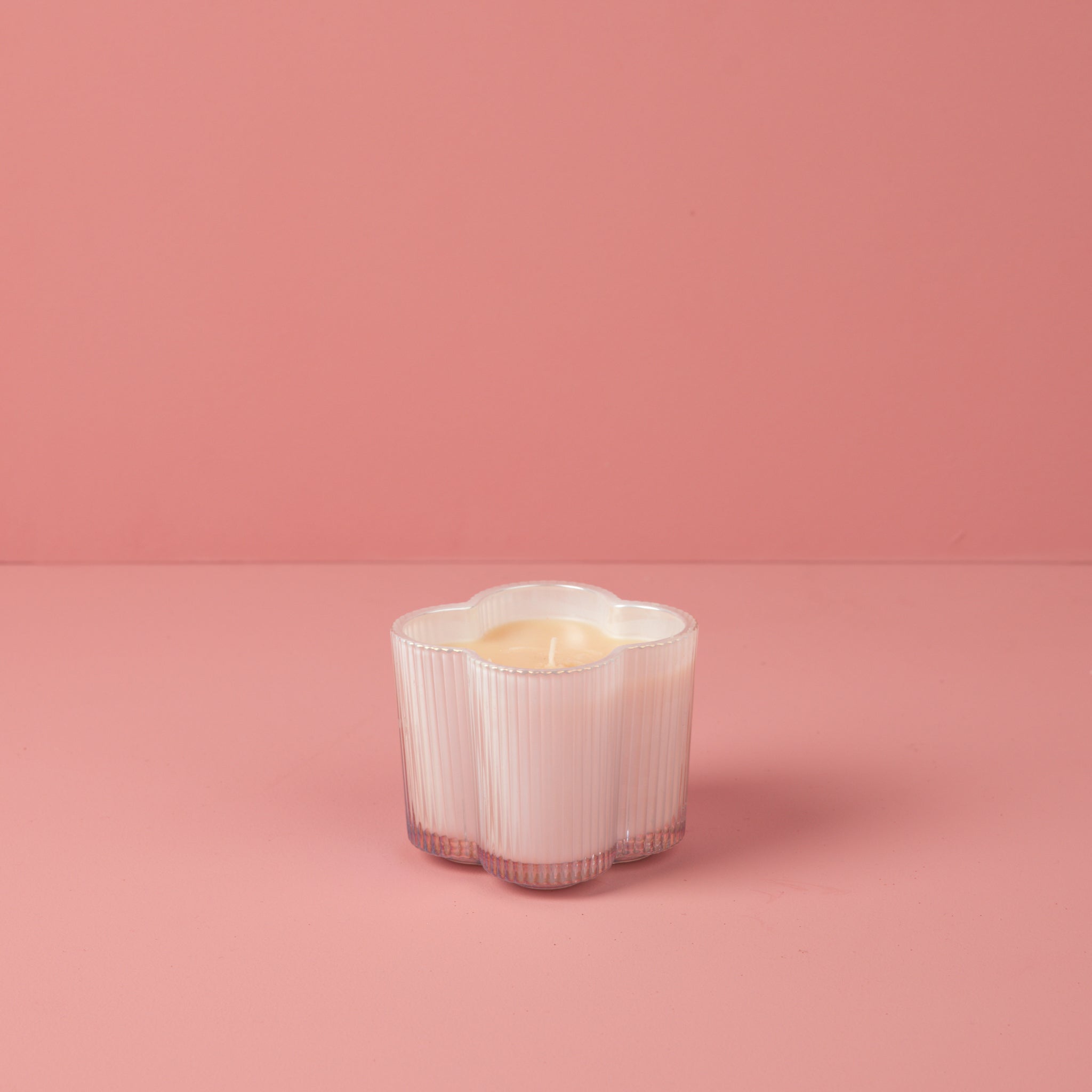 The Sweet Grace Collection Candle #076 by Bridgewater Candle is a small, white, flower-shaped soy-blend candle in a ribbed glass holder displayed on a pink surface against a pink background.