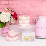 A pastel pink kitchen scene features a pink electric kettle, white and red flowers in a pink vase, the Sweet Grace Collection Candle #074 by Bridgewater Candle, a pink cup and saucer, and an English tea tin against a pink brick wall.