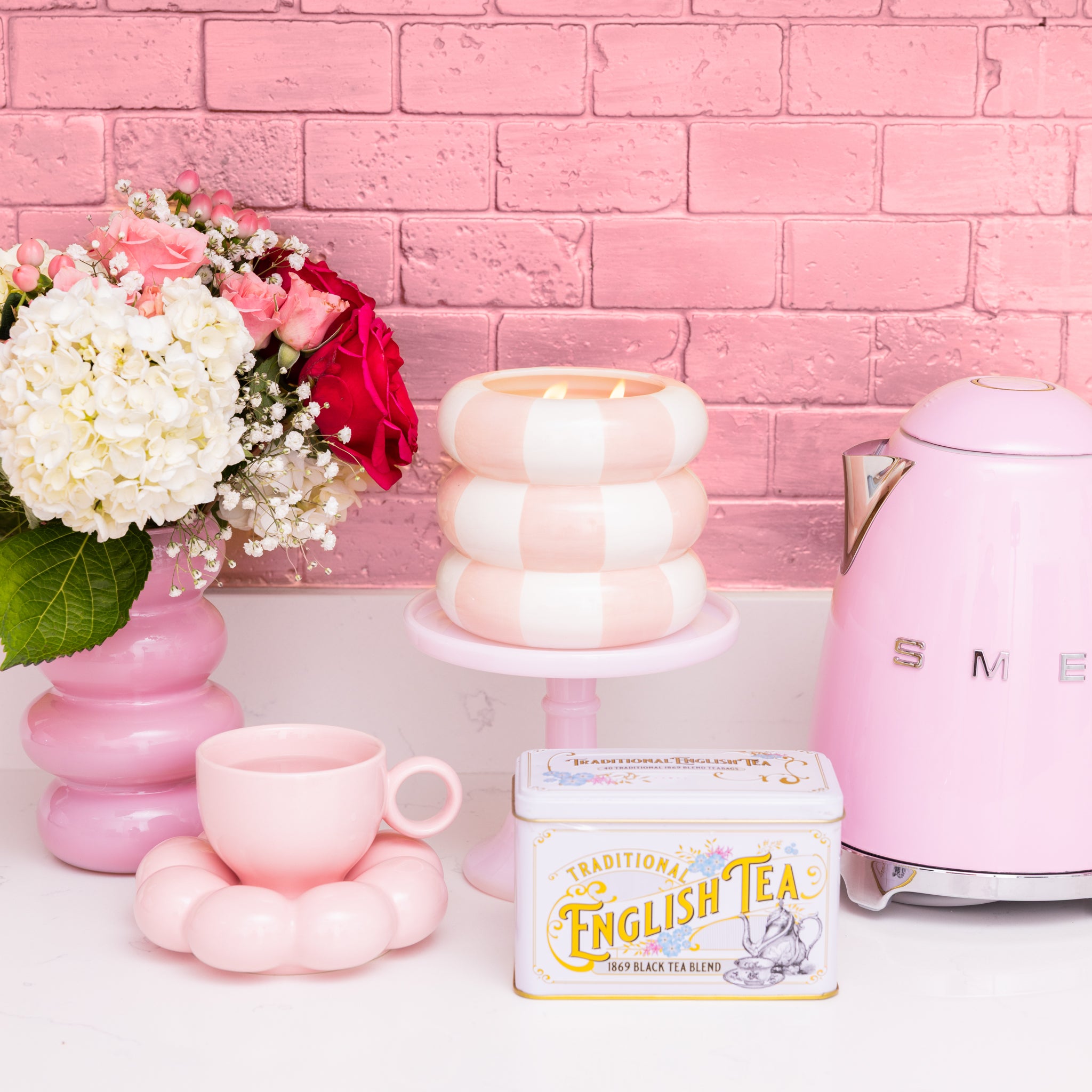 A pastel pink kitchen scene features a pink electric kettle, white and red flowers in a pink vase, the Sweet Grace Collection Candle #074 by Bridgewater Candle, a pink cup and saucer, and an English tea tin against a pink brick wall.
