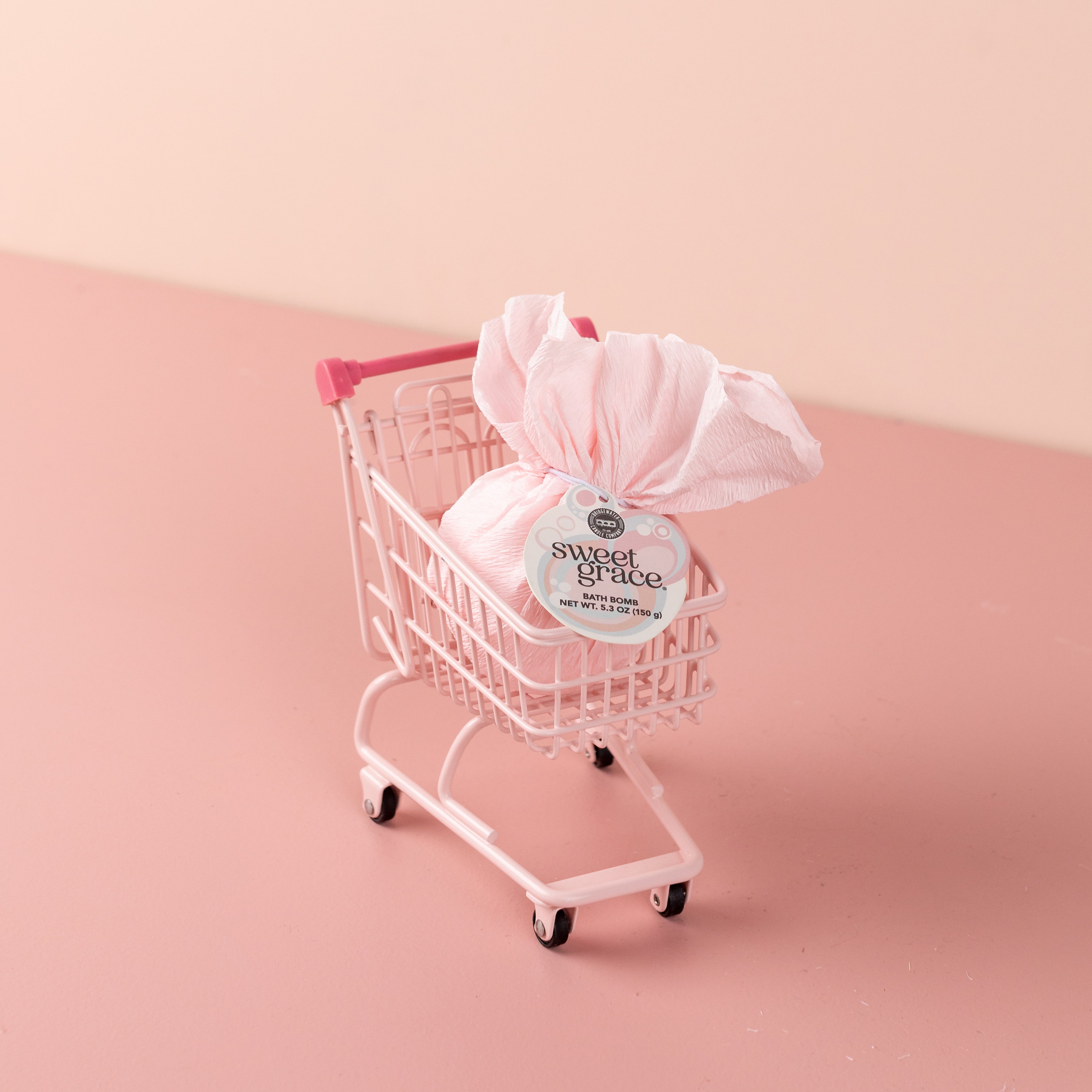 A small pink shopping cart holds a wrapped Bath Bomb 4 Pack-Sweet Grace by Bridgewater Candle, set against a matching pink background.