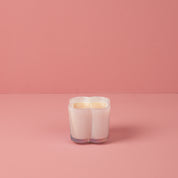 The Bridgewater Candle Sweet Grace Collection Candle #076 is a white, heart-shaped soy-blend candle in a translucent holder, displayed on a pink surface with a matching pink background.