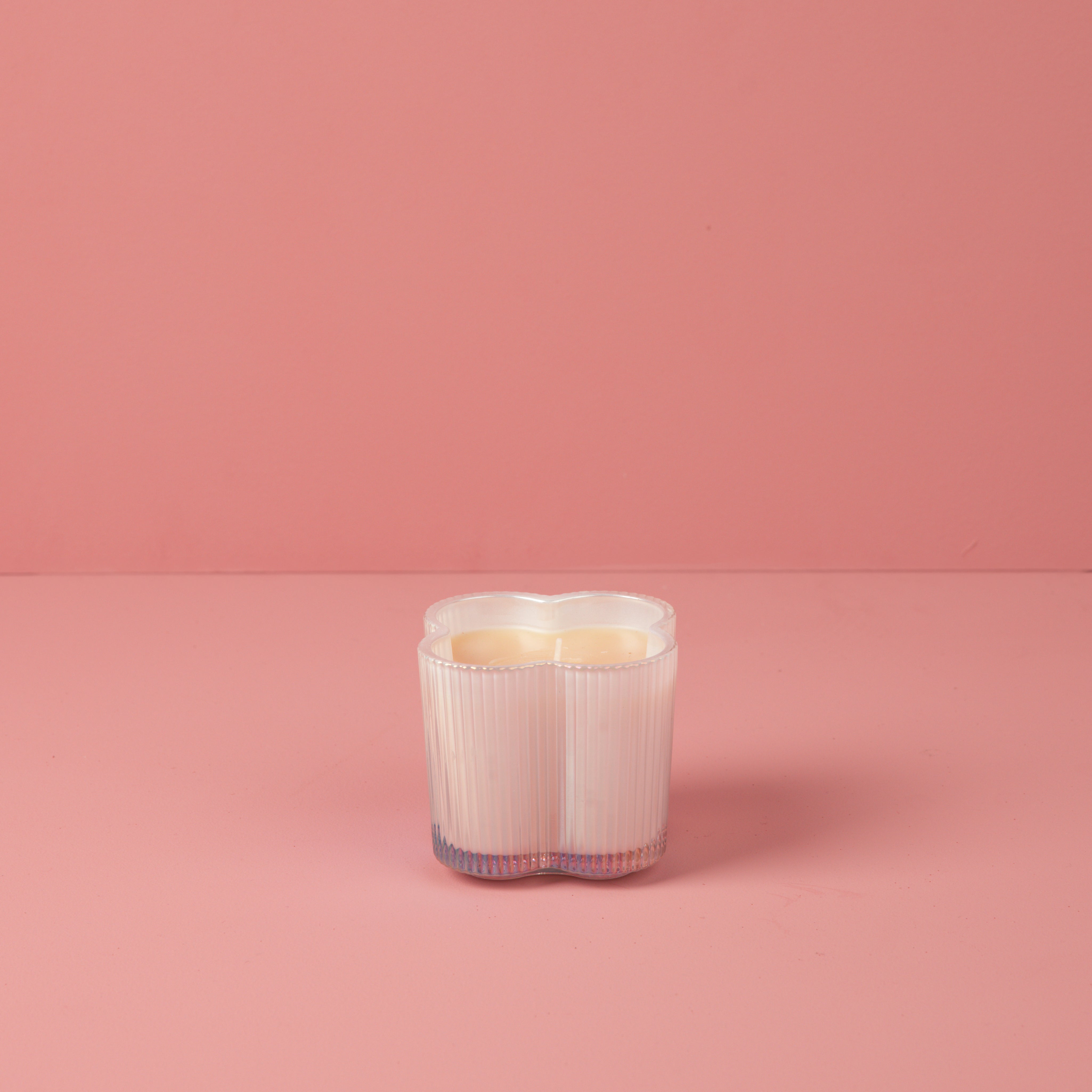 The Bridgewater Candle Sweet Grace Collection Candle #076 is a white, heart-shaped soy-blend candle in a translucent holder, displayed on a pink surface with a matching pink background.