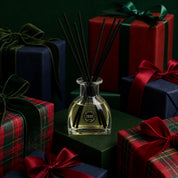 The I'll Be Home Reed Diffuser by Bridgewater Candle, a glass diffuser with black fiber reeds, sits amid dark green, red, and plaid-wrapped gifts tied with velvet ribbons against a dark green backdrop.