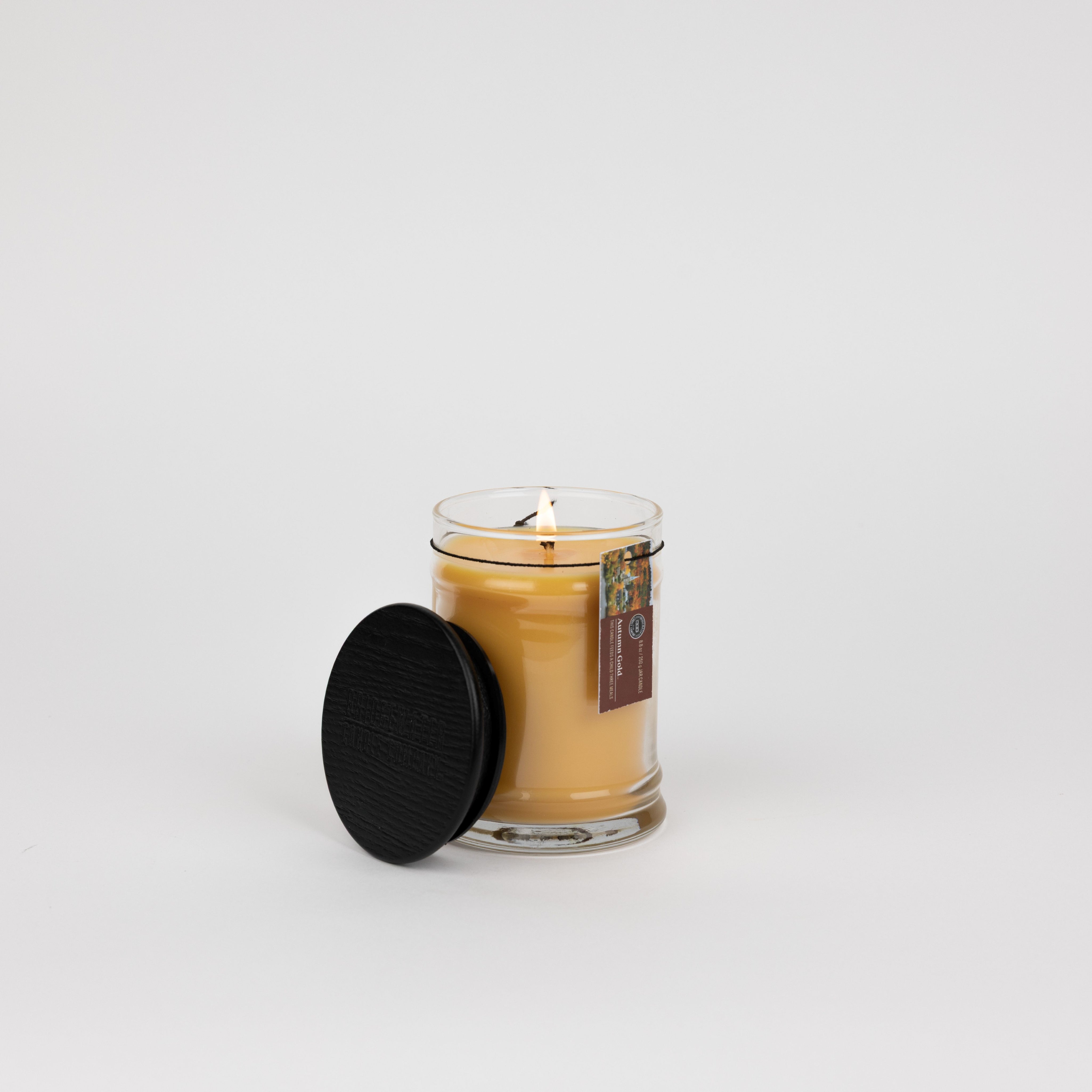 The Bridgewater Candle Autumn Gold Small Jar Candle, made with soy-blend wax and a warm fall scent, sits in a glass jar with its black textured lid beside it on a white background. A small product label accents the jar.