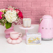 A pastel pink kitchen with a pink kettle, English tea tin, pink cup on a flower coaster, two cups on a cake stand, white and pink flowers in a vase, and Bridgewater Candle’s Sweet Grace Collection Candle #076 against the pink brick wall.