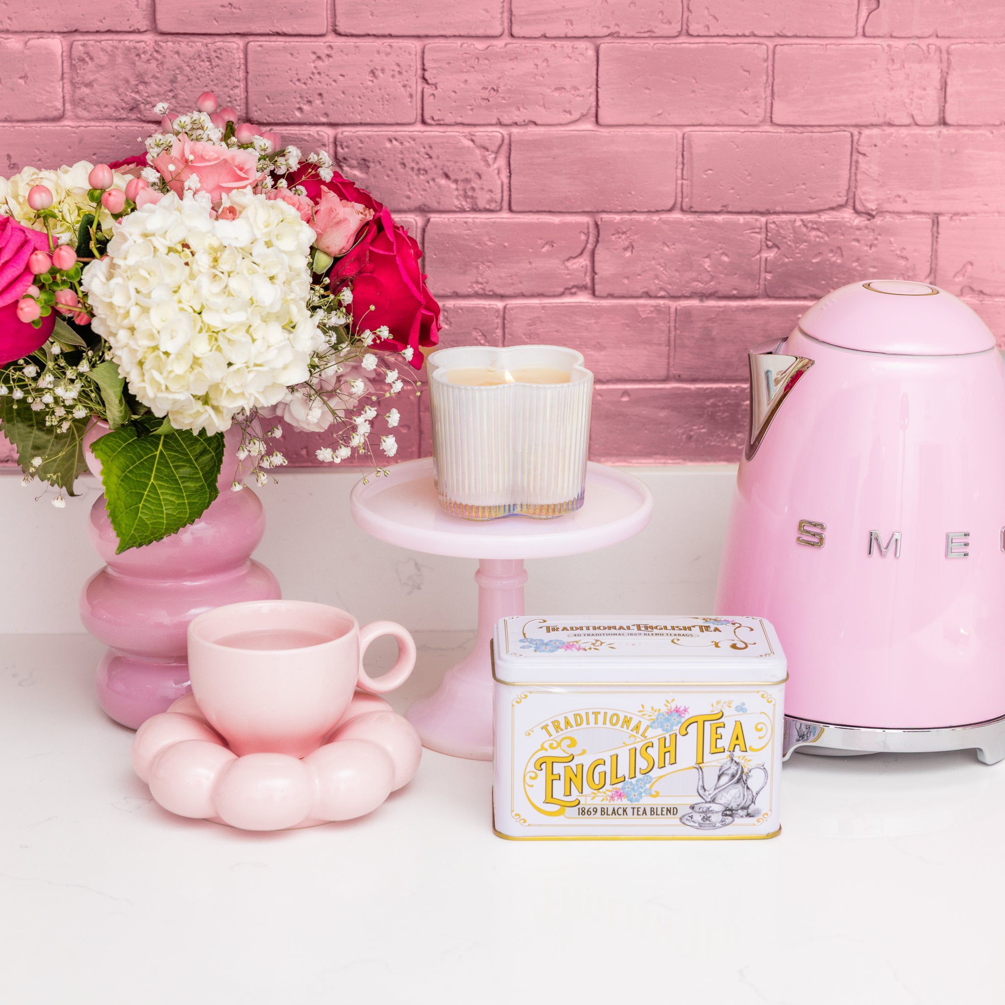 A pastel pink kitchen with a pink kettle, English tea tin, pink cup on a flower coaster, two cups on a cake stand, white and pink flowers in a vase, and Bridgewater Candle’s Sweet Grace Collection Candle #076 against the pink brick wall.