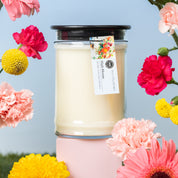 The Bridgewater Candle High Bloom 18oz Large Jar Candle, made with soy-blend wax in a glass jar with a black lid and floral tag, is surrounded by vibrant pink, yellow, and red flowers on a light blue background.