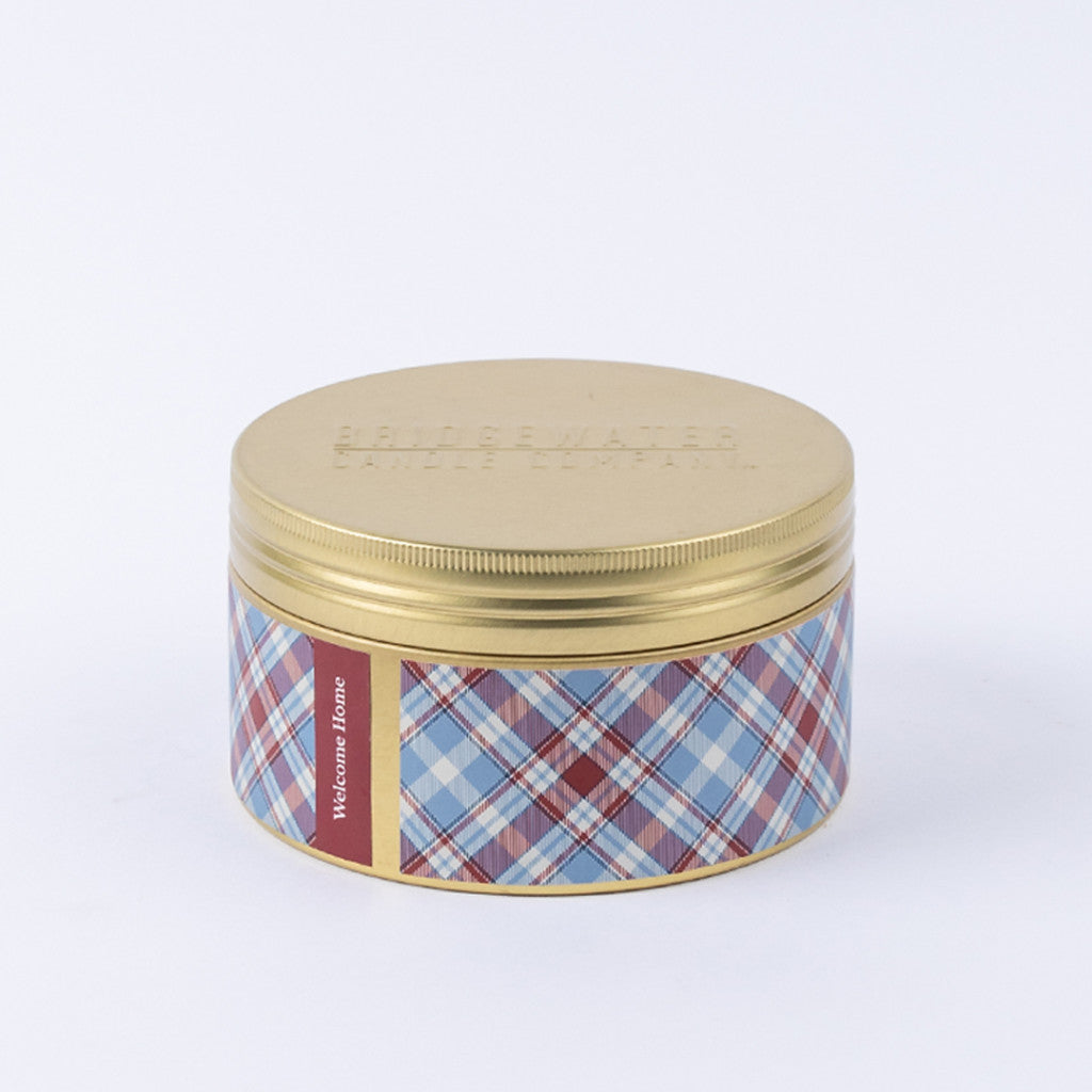 The Bridgewater Candle Company Welcome Home 3-Wick Tin Candle features a gold lid and blue, red, and white plaid-patterned label with “Welcome Home” text. It’s filled with soy-blend wax for an inviting touch.