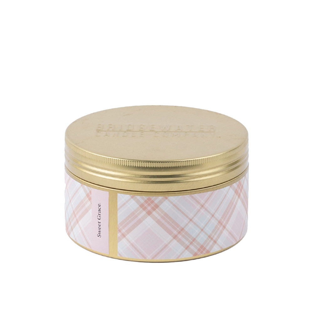 The Bridgewater Candle Company Sweet Grace 3-Wick Tin Candle features a gold lid, plaid design in pink, white, and beige, and a “Sweet Grace” label. Crafted from soy-blend wax for an elegant, fragrant accent to your home.