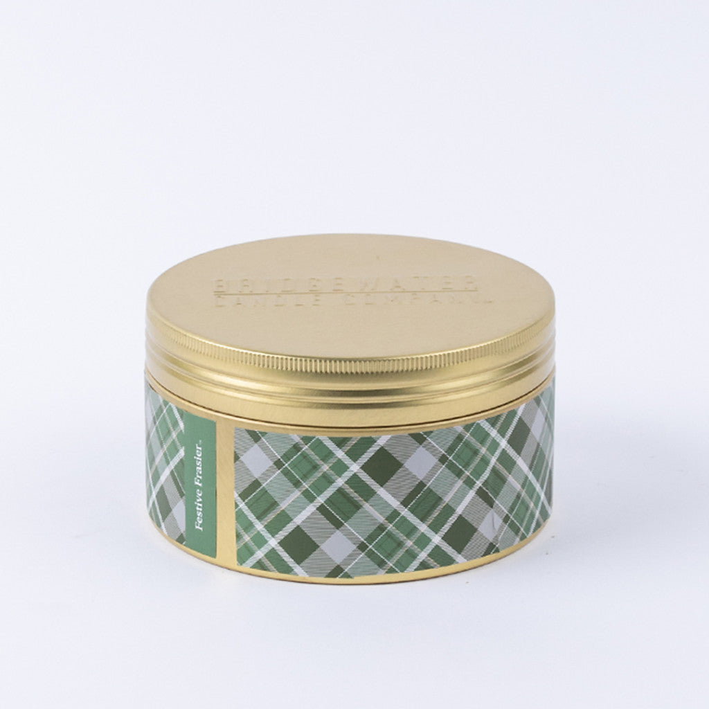 A round gold tin from Bridgewater Candle Company, labeled "Festive Frasier Holiday 3-Wick Tin Candle" in green plaid, features soy-blend wax and is shown against a plain white background.