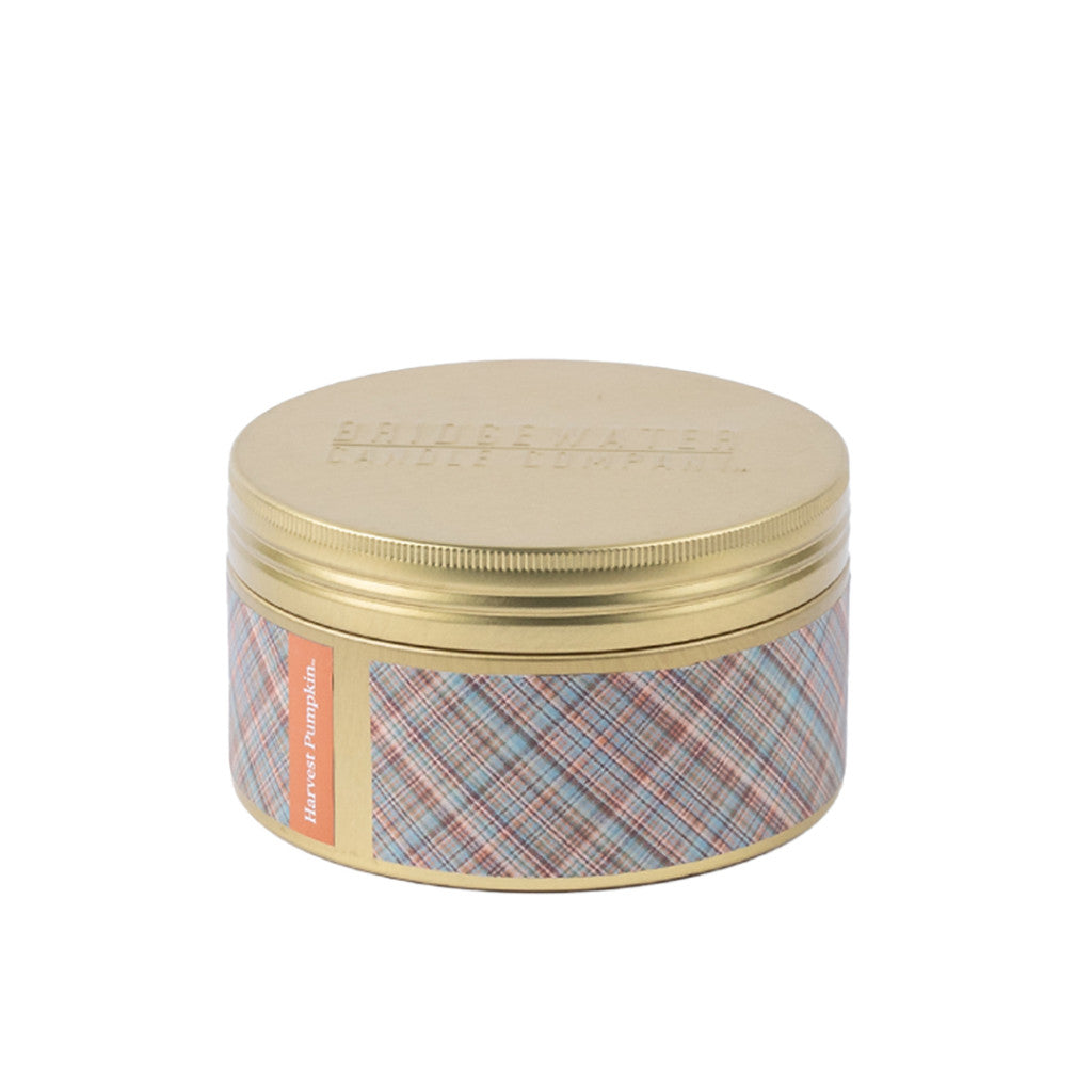 A round, gold metal tin from Bridgewater Candle Company features a plaid-patterned label and an orange tag that reads “Harvest Pumpkin.” The lid is embossed with “BRIDGEWATER,” and this inviting Harvest Pumpkin 3-Wick Tin Candle offers a warm pumpkin spice fragrance.