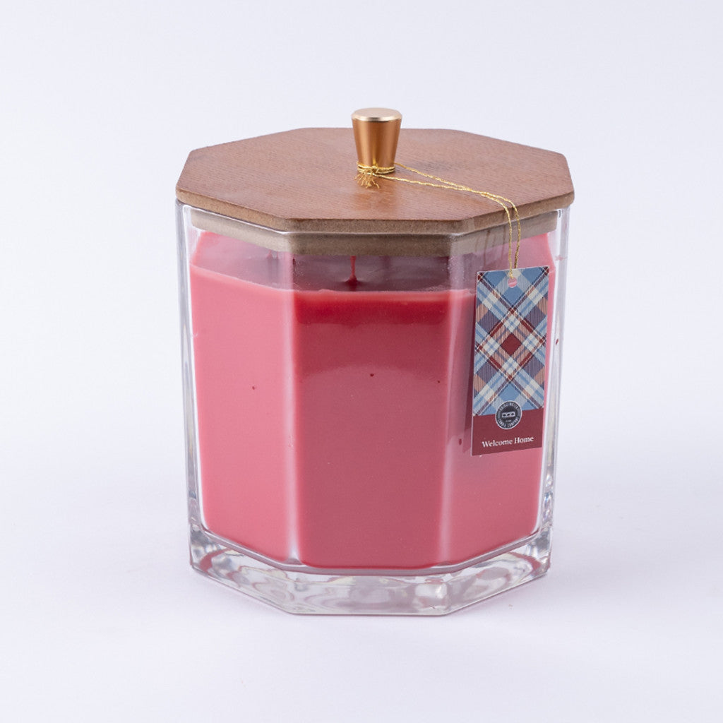 The Bridgewater Candle Company Welcome Home Large Decorative Candle features pink soy-blend wax in a clear hexagonal glass jar with a wooden lid and gold knob, plus a plaid tag attached with gold string—perfect for home decor.