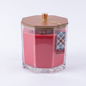 The Bridgewater Candle Company Welcome Home Large Decorative Candle features pink soy-blend wax in a clear hexagonal glass jar with a wooden lid and gold knob, plus a plaid tag attached with gold string—perfect for home decor.