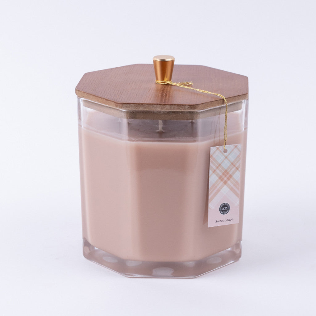 The Bridgewater Candle Company Sweet Grace Large Decorative Candle features beige soy-blend wax in a hexagonal glass jar with a wooden lid and gold knob. A plaid tag is attached with a gold string, all shown on a plain white background.