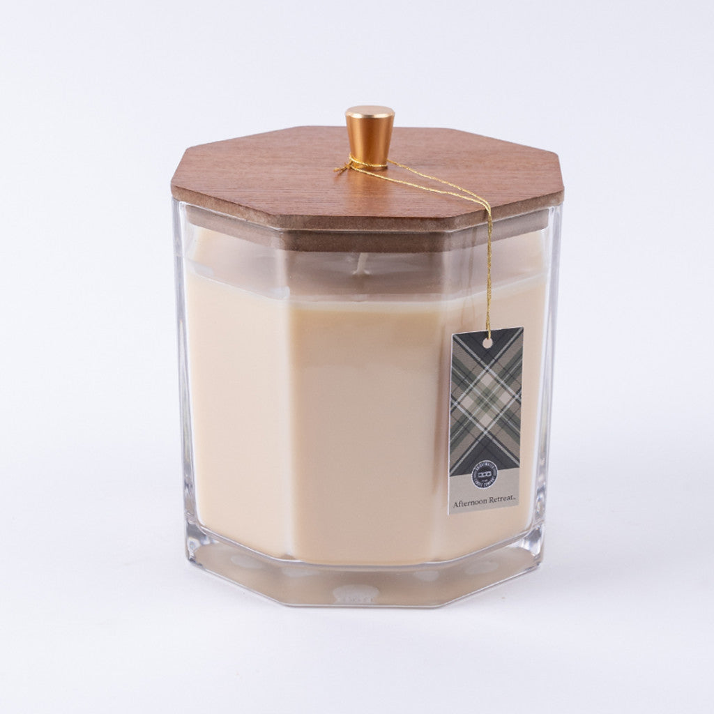 The Afternoon Retreat Large Decorative Candle by Bridgewater Candle Company features cream soy-blend wax in a hexagonal glass jar with a wooden lid, gold knob, string, and plaid tag labeled Afternoon Bonfire. Background is white.