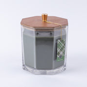 The Bridgewater Candle Company Festive Frasier Holiday Large Decorative Candle features soy-blend wax in a clear hexagonal jar with a wooden lid, gold knob, and green plaid tag tied with gold string. Background is white.