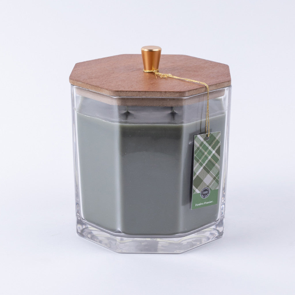 The Bridgewater Candle Company Festive Frasier Holiday Large Decorative Candle features soy-blend wax in a clear hexagonal jar with a wooden lid, gold knob, and green plaid tag tied with gold string. Background is white.