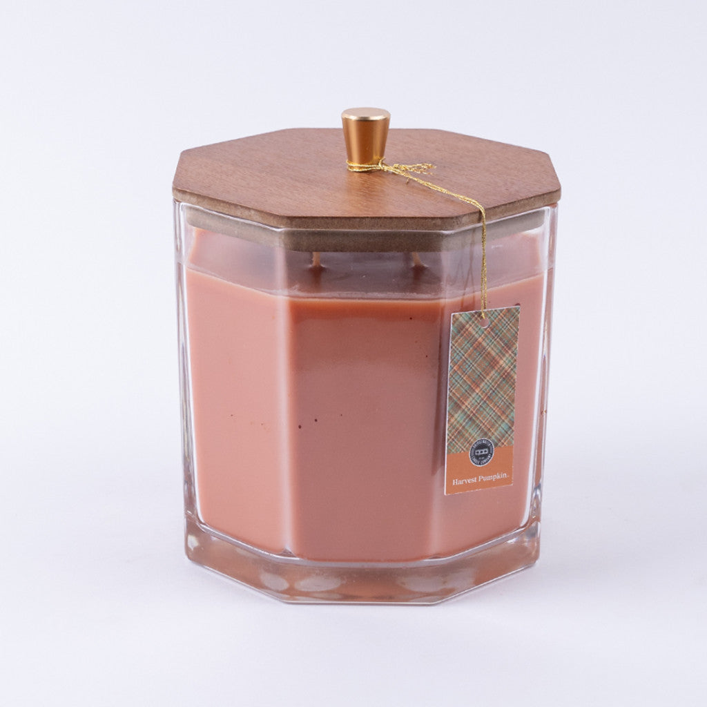 The Bridgewater Candle Company Harvest Pumpkin Holiday Large Decorative Candle features orange soy-blend wax in a hexagonal glass jar with a wooden lid and gold knob, plus a plaid tag reading "Harvest Pumpkin," all set against a plain white background.