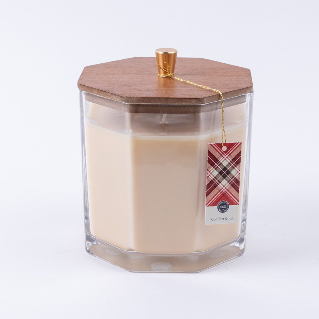 The Bridgewater Candle Company’s Comfort & Joy Holiday Large Decorative Candle features cream soy-blend wax in a hexagonal glass jar with a wooden lid, gold knob, plaid tag, and premium fragrance, set against a white background.