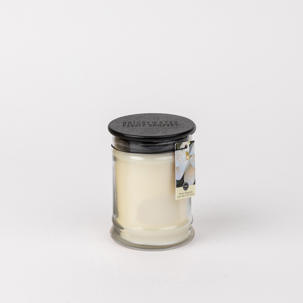 The Bridgewater Candle Company Sweet Magnolia 8oz Jar Candle comes in a small clear glass with a black lid and floral tag, ideal for infusing any space with a delightful magnolia fragrance.