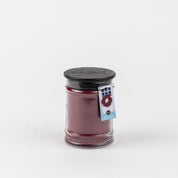 The Bridgewater Candle Company Welcome Home 8oz Jar Candle features deep reddish-purple soy-blend wax in a large glass jar with a dark wooden lid and a wreath tag, shown against a plain white background.