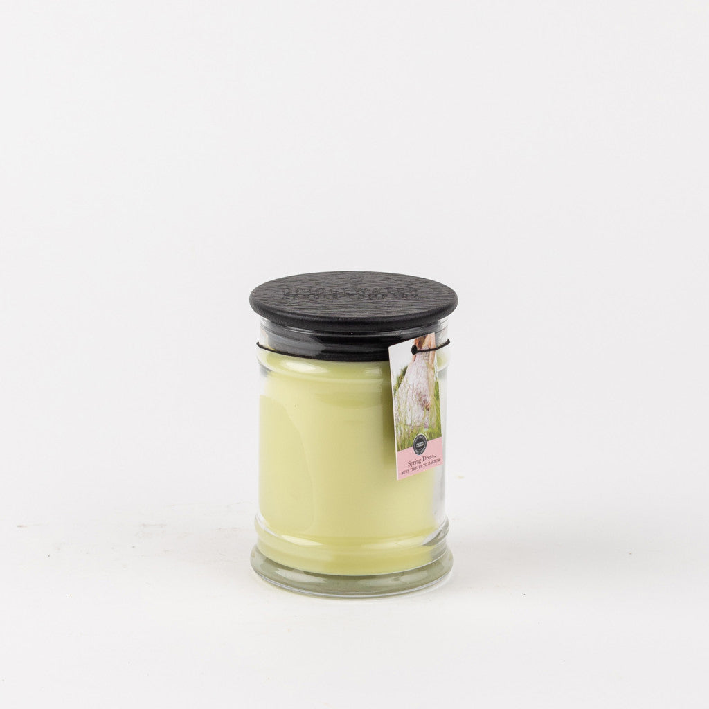 The Bridgewater Candle Company Spring Dress 8oz Jar Candle features pale yellow wax in a small jar with a black lid and a pink-labeled tag. Set against a white background, it’s perfect for enjoying a delicate floral scent.