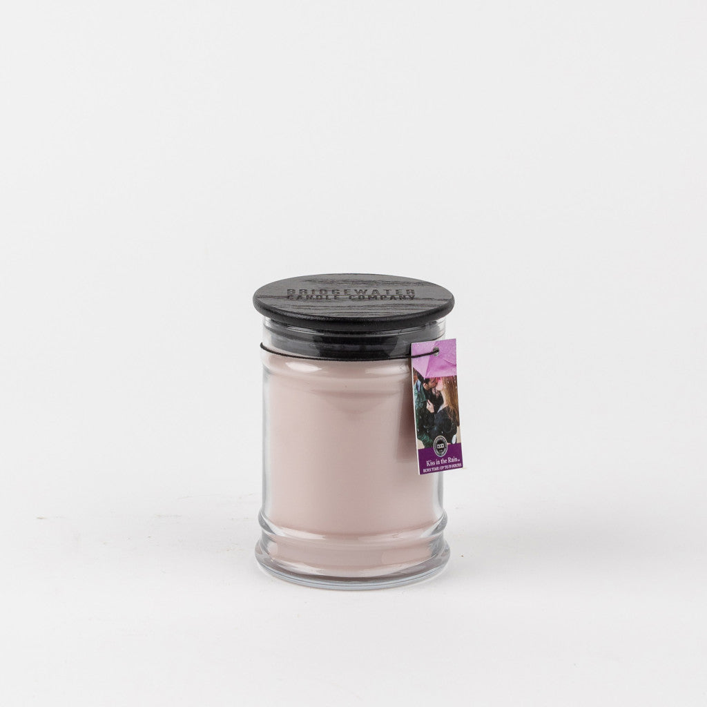 A Bridgewater Candle Company Kiss In The Rain 8oz Jar Candle with a black lid and a small tag, filled with pale pink wax, is displayed against a plain white background.