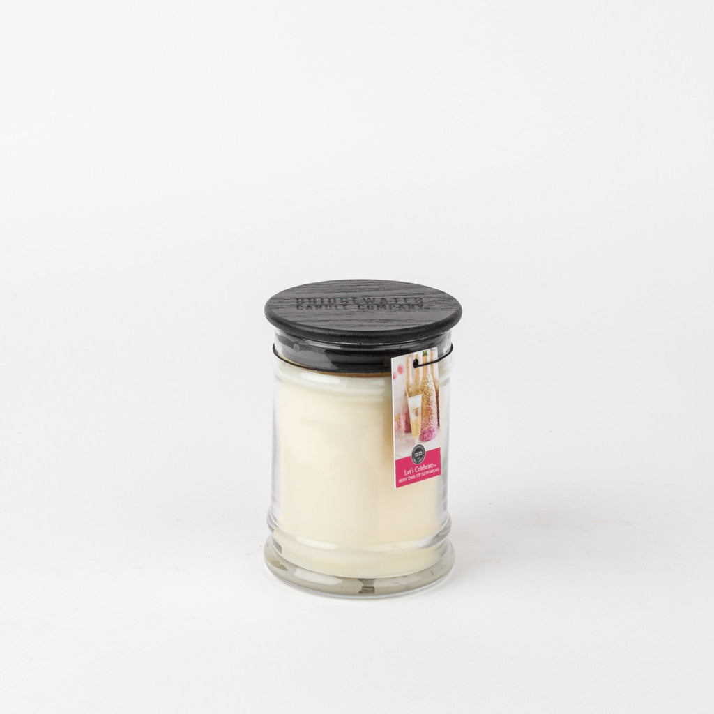 A Bridgewater Candle Company Lets Celebrate 8oz Jar Candle with a black lid and a label featuring a drink and LIGHT A CANDLE FEED A CHILD, displayed against a plain white background.