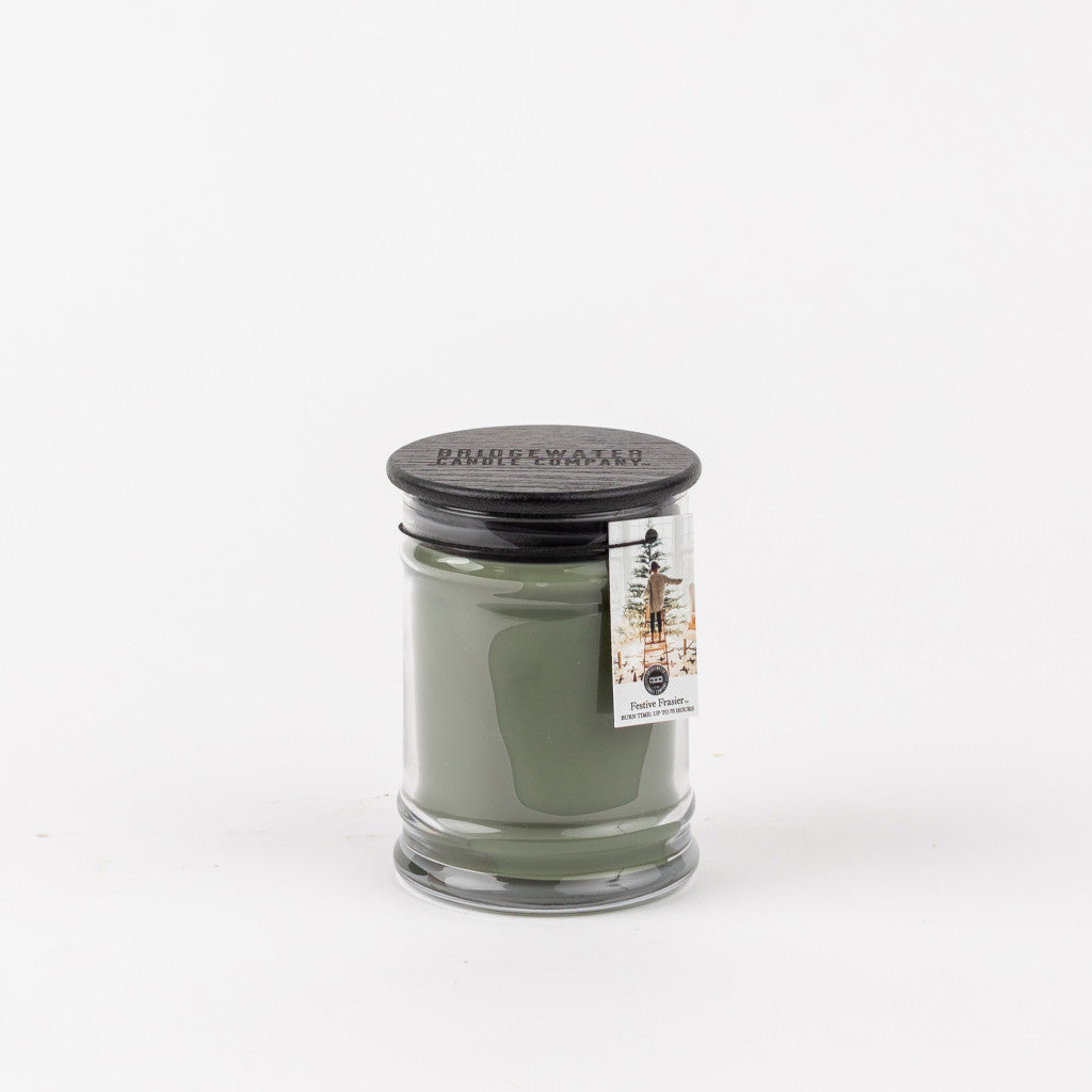 A green Festive Frasier 8oz Jar Candle by Bridgewater Candle Company in a clear glass jar with a black lid. A small tag featuring a tree illustration and Festive Frasier text adds charm, set against a plain white background.
