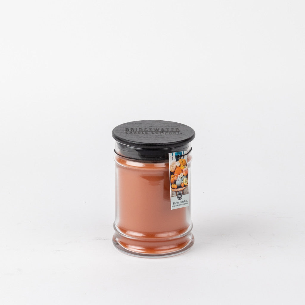 The Harvest Pumpkin 8oz Jar Candle by Bridgewater Candle Company features orange wax, a black lid, and a label with pumpkins and baked goods. This cozy autumn scent adds fall ambiance to any room. Shown against a plain white background.